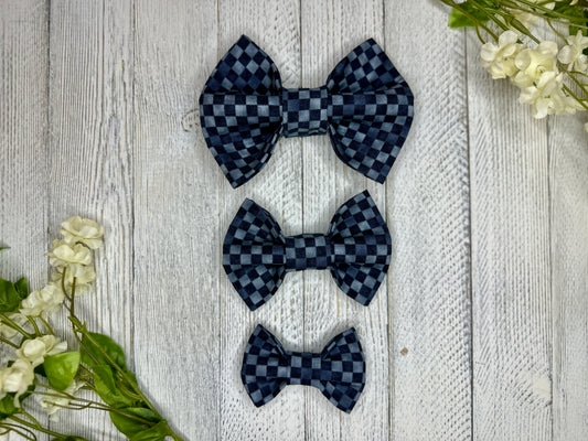 Three pet bow ties (small, medium, large) with a black and grey checkered pattern, displayed on a wooden surface with flowers for decoration. 