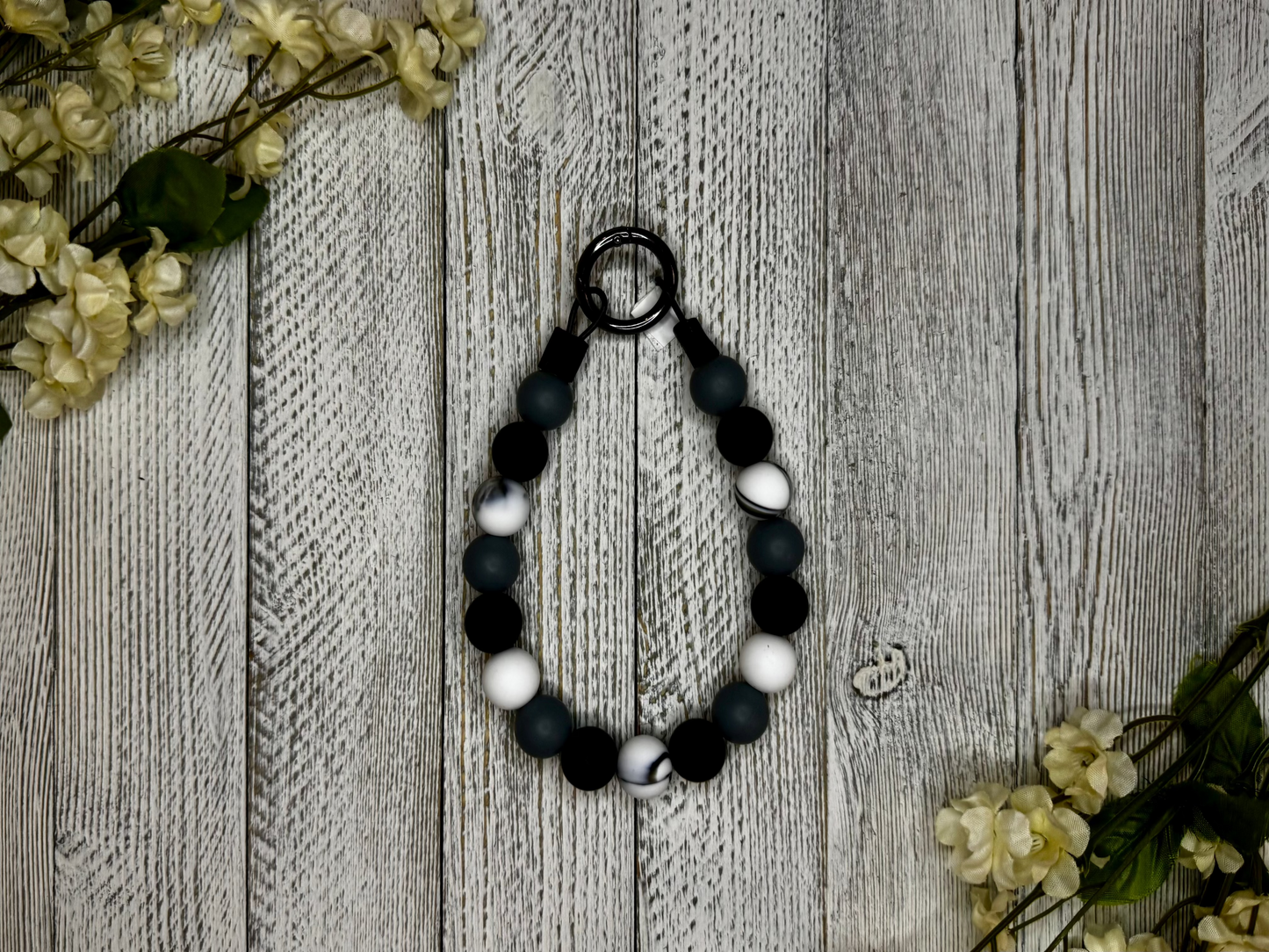 Beaded pet necklace with a gray, black, and white pattern. Displayed on a wooden surface with flowers for decoration. 