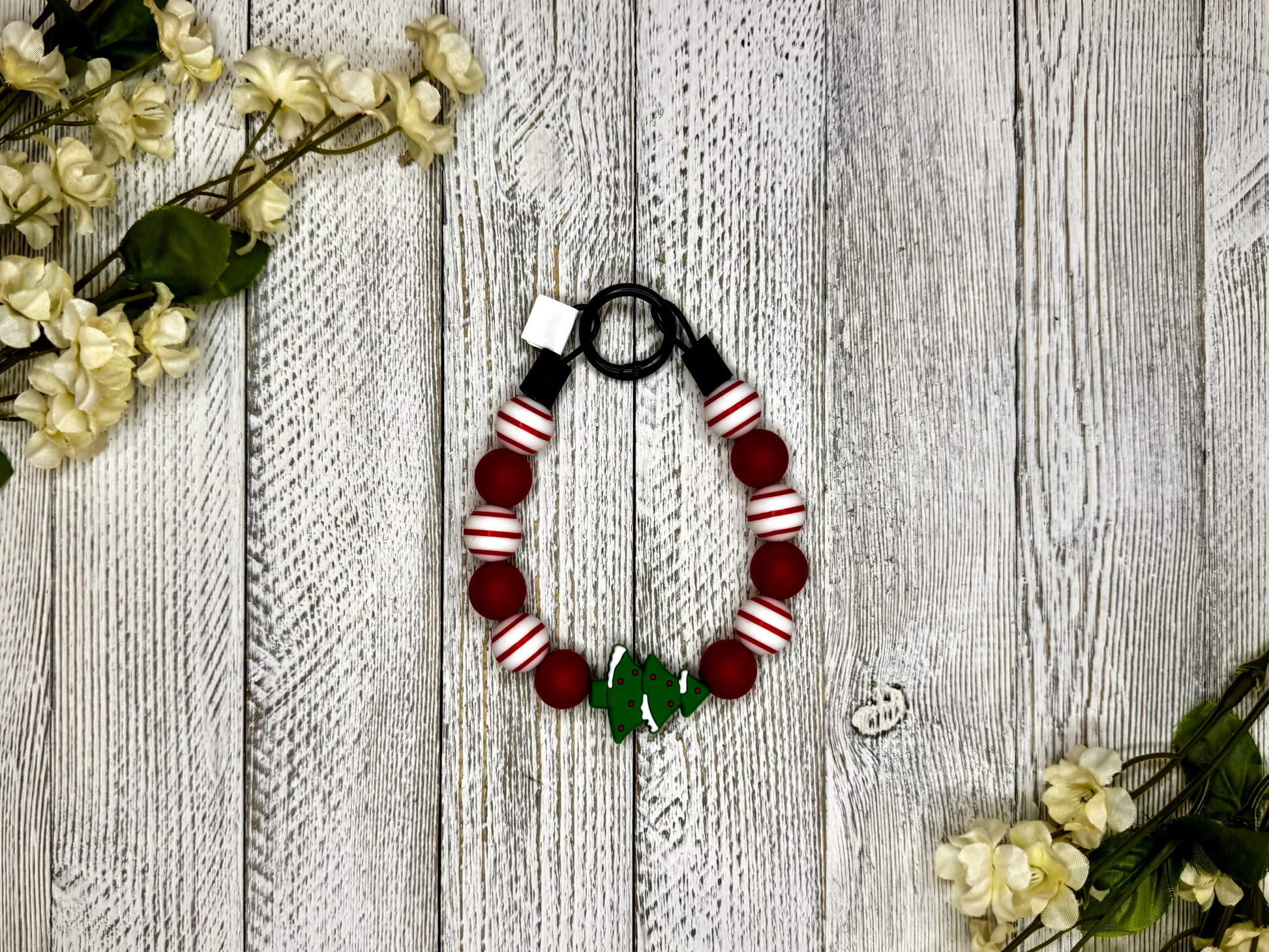 Beaded pet necklace with a red and white pattern, including a Christmas tree. Displayed on a wooden surface with flowers for decoration. 