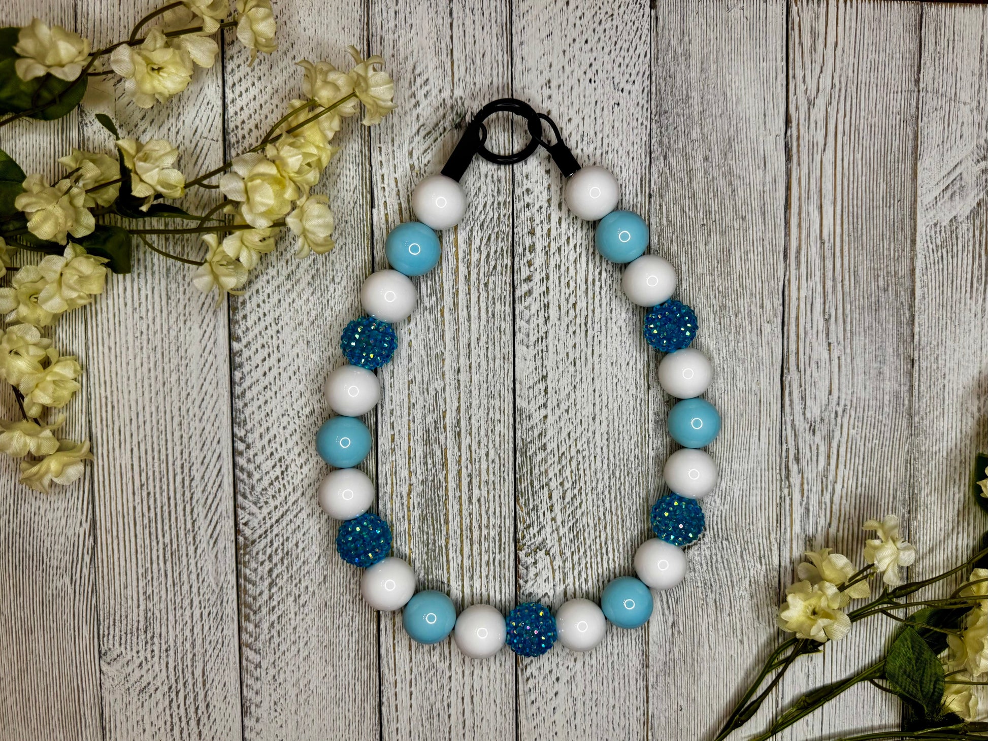 Beaded dog necklace with a light blue and white pattern with sparkles. Displayed on a wooden surface with flowers for decoration.