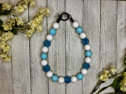 Beaded dog necklace with a light blue and white pattern with sparkles. Displayed on a wooden surface with flowers for decoration.