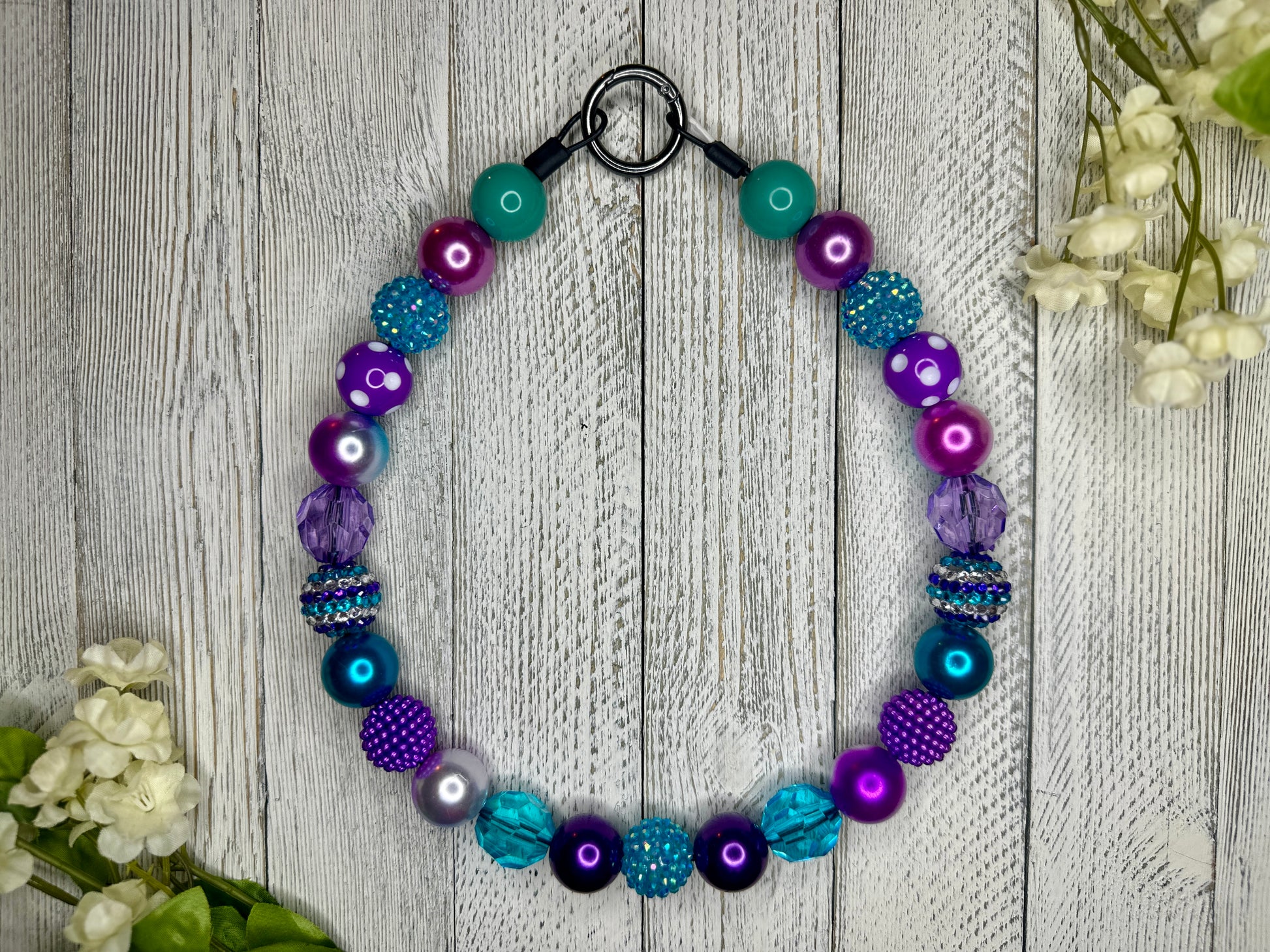 Beaded pet necklace with a purple and teal pattern, including polka dots. Displayed on a wooden surface with flowers for decoration. 