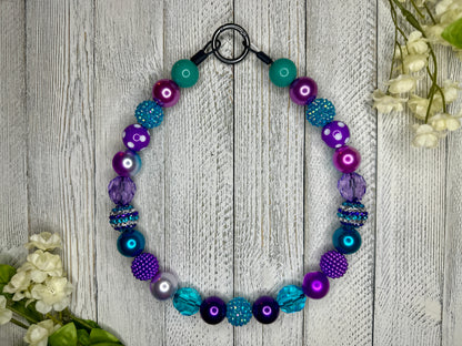 Beaded pet necklace with a purple and teal pattern, including polka dots. Displayed on a wooden surface with flowers for decoration. 