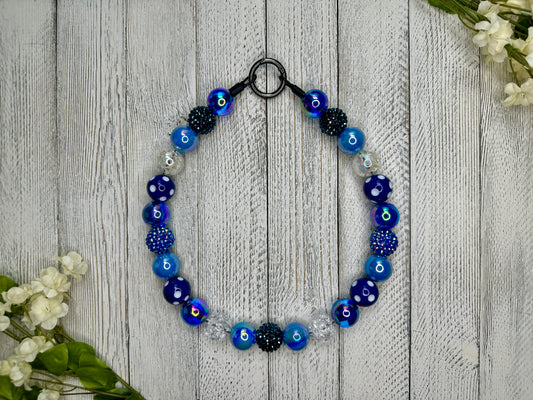 Beaded pet necklace with a navy blue pattern including sparkles and polka dots. Displayed on a wooden surface with flowers for decoration.