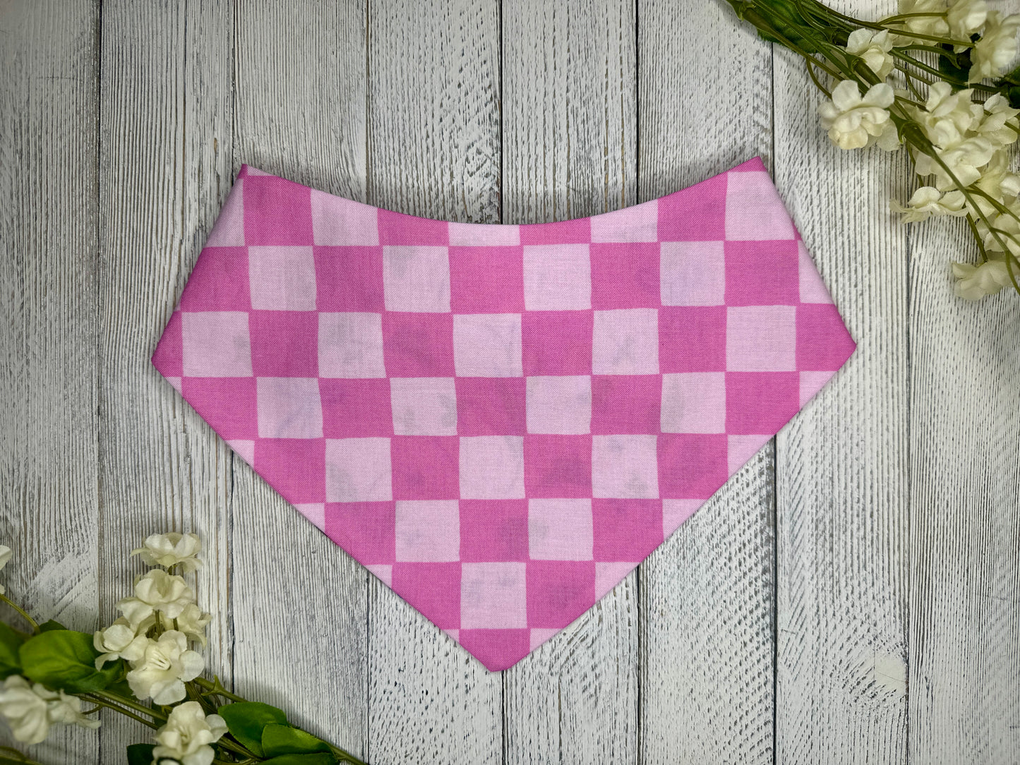 A pet bandana with a light and dark pink checkered pattern, displayed on a wooden surface with flowers for decoration. 