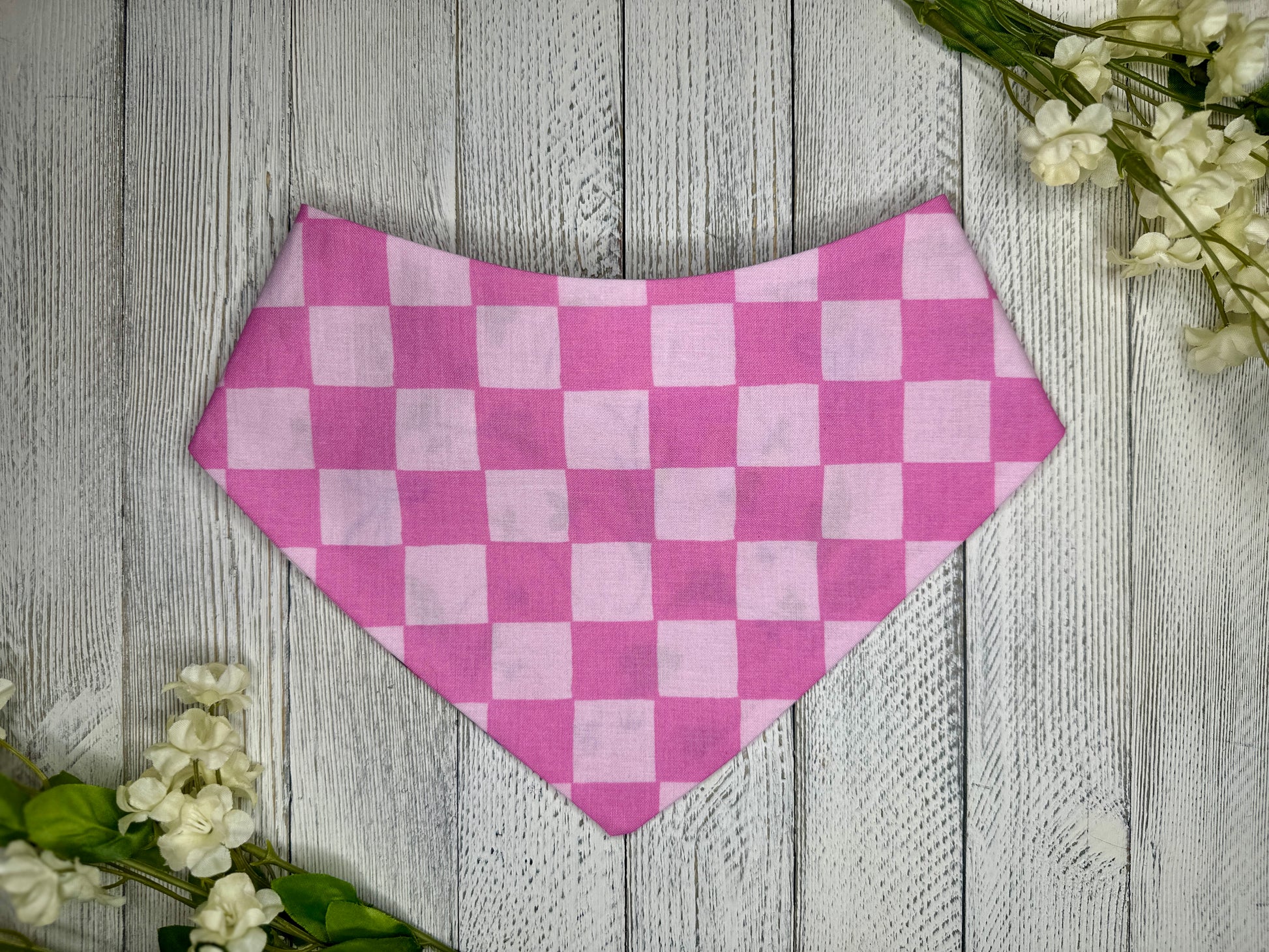 A pet bandana with a light and dark pink checkered pattern, displayed on a wooden surface with flowers for decoration. 