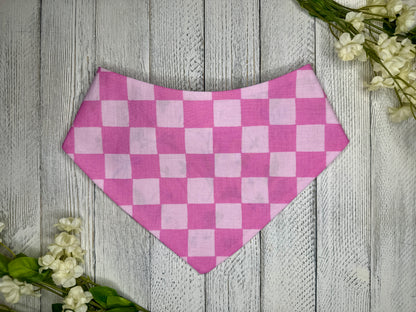 A pet bandana with a light and dark pink checkered pattern, displayed on a wooden surface with flowers for decoration. 