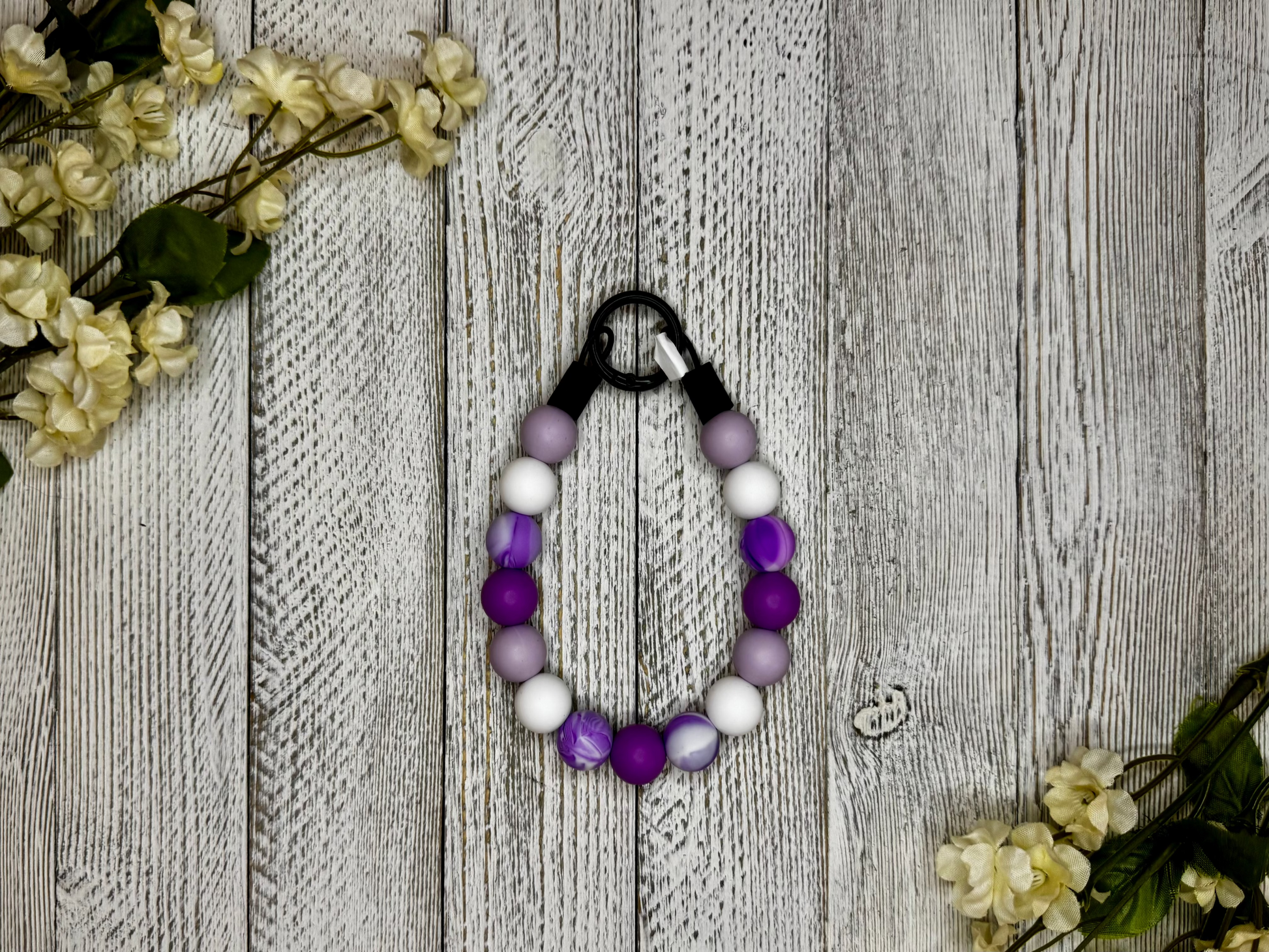 Beaded pet necklace with a purple and white pattern, including marbling. Displayed on a wooden surface with flowers for decoration. 