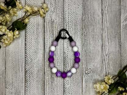 Beaded pet necklace with a purple and white pattern, including marbling. Displayed on a wooden surface with flowers for decoration. 