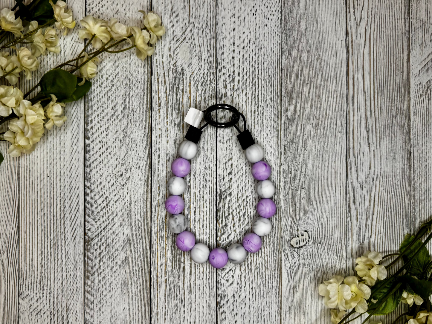 Beaded pet necklace with a purple and white pattern, including marbling. Displayed on a wooden surface with flowers for decoration. 