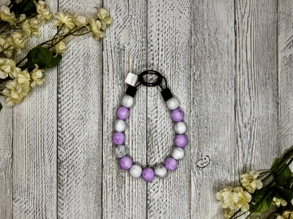 Beaded pet necklace with a purple and white pattern, including marbling. Displayed on a wooden surface with flowers for decoration. 