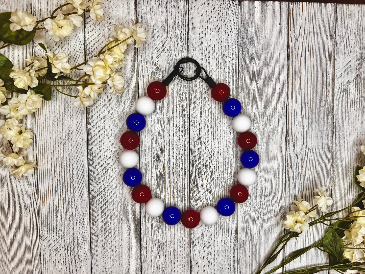 Beaded pet necklace with a red, white, and blue pattern. Displayed on a wooden surface with flowers for decoration. 