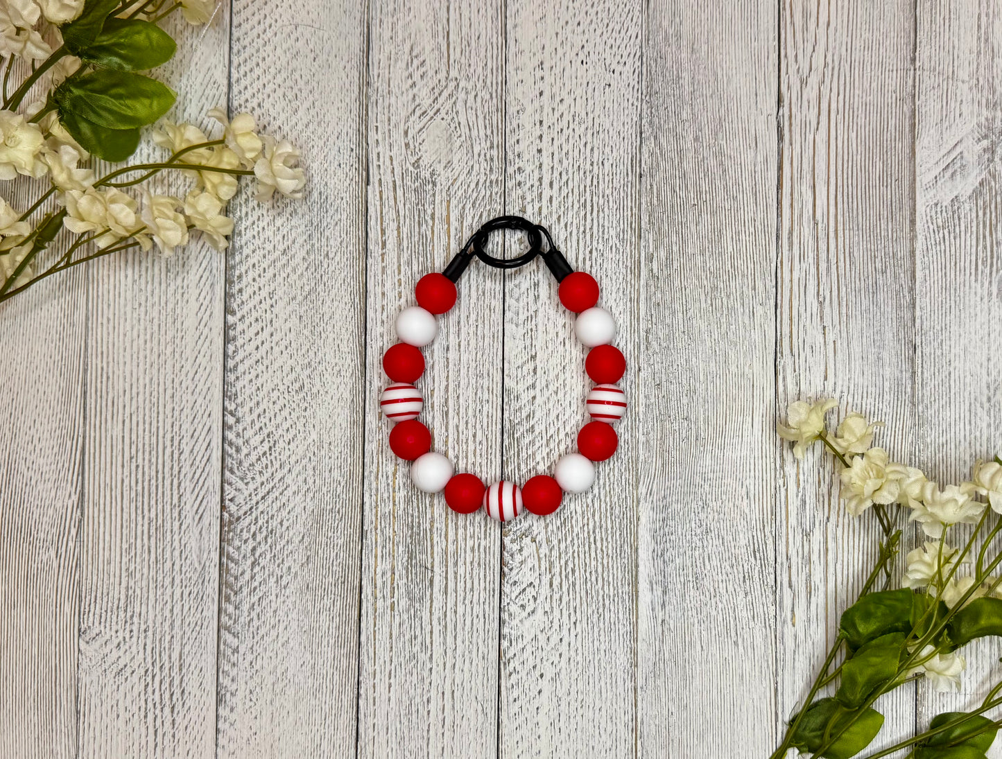 Beaded pet necklace with a red and white pattern, including stripes. Displayed on a wooden surface with flowers for decoration. 