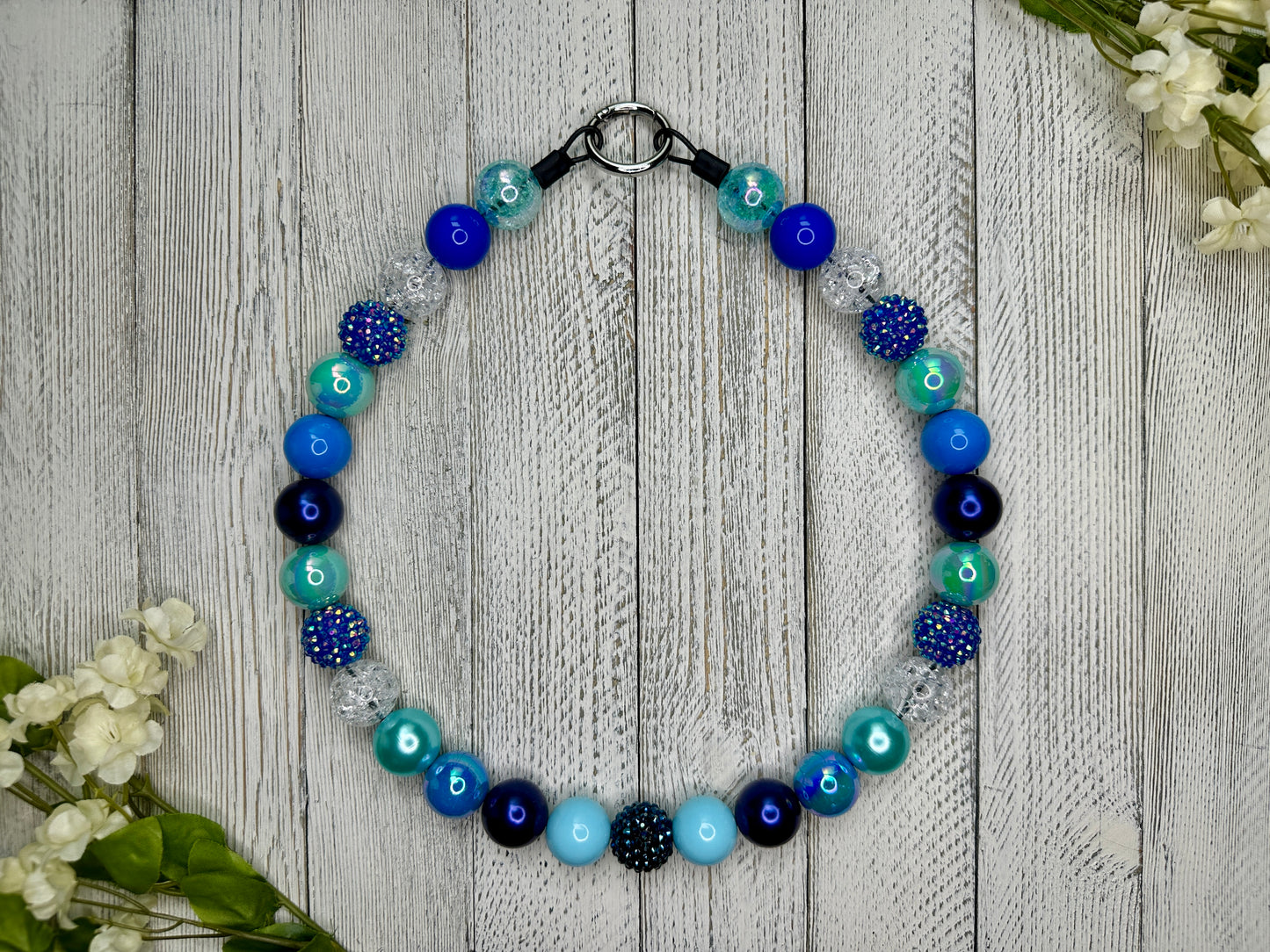 Beaded pet necklace with a variety of blues pattern, including sparkles. Displayed on a wooden surface with flowers for decoration. 
