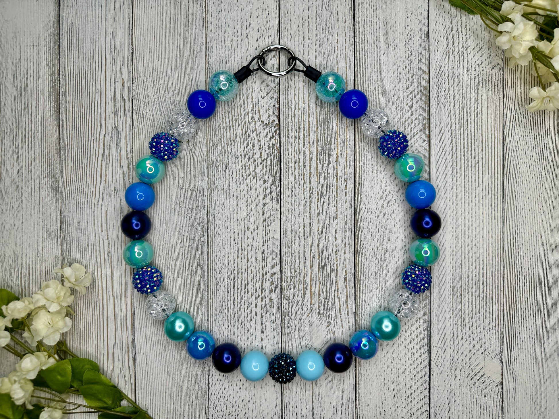 Beaded pet necklace with a variety of blues pattern, including sparkles. Displayed on a wooden surface with flowers for decoration. 