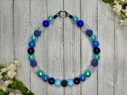 Beaded pet necklace with a variety of blues pattern, including sparkles. Displayed on a wooden surface with flowers for decoration. 