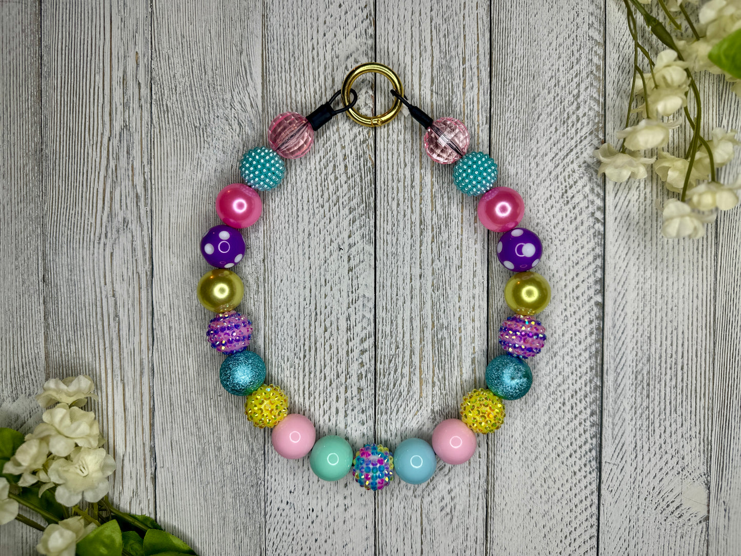 Beaded pet necklace with a spring pattern, including sparkles. Displayed on a wooden surface with flowers for decoration. 