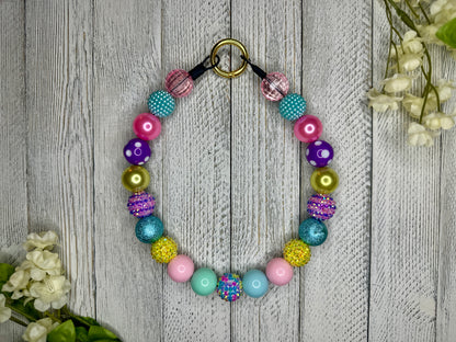 Beaded pet necklace with a spring pattern, including sparkles. Displayed on a wooden surface with flowers for decoration. 