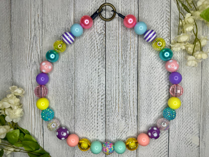 Beaded pet necklace with a spring pattern, including polka dots and stripes. Displayed on a wooden surface with flowers for decoration. 