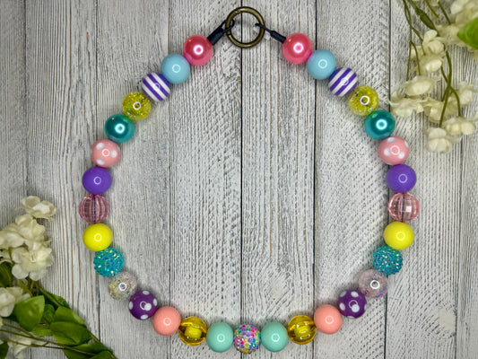 Beaded pet necklace with a spring pattern, including polka dots and stripes. Displayed on a wooden surface with flowers for decoration. 