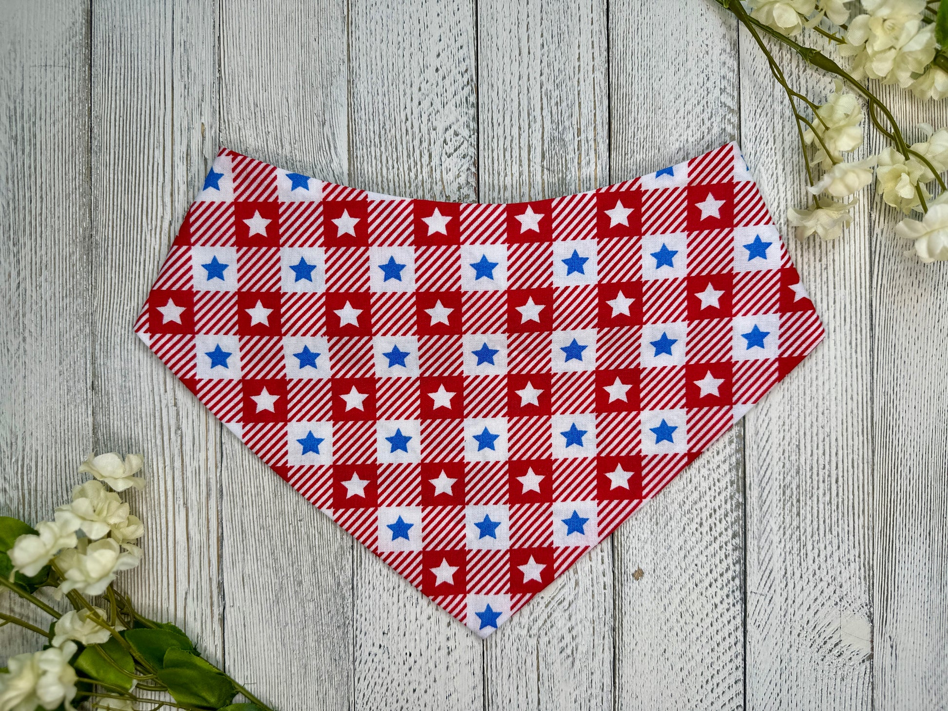 A pet bandana with a red and white checkered pattern with stars, displayed on a wooden surface with flowers for decoration. 