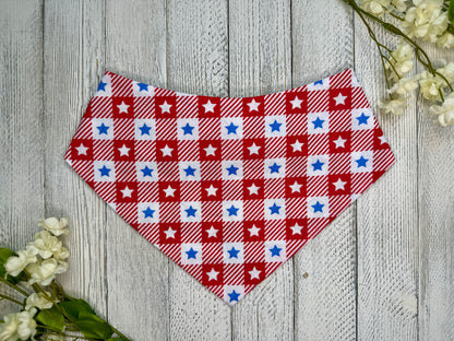 A pet bandana with a red and white checkered pattern with stars, displayed on a wooden surface with flowers for decoration. 