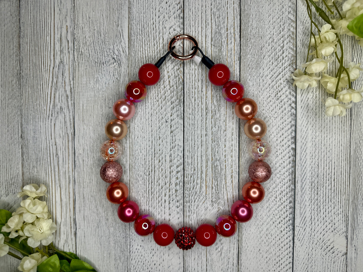 Beaded pet necklace with a red sunset pattern, including sparkles. Displayed on a wooden surface with flowers for decoration. 