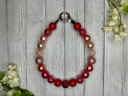 Beaded pet necklace with a red sunset pattern, including sparkles. Displayed on a wooden surface with flowers for decoration. 