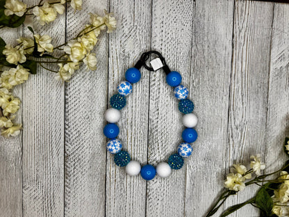 Beaded dog necklace with a blue and white pattern including snowflakes. Displayed on a wooden surface with flowers for decoration.